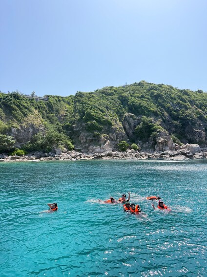 A group of people swimming in a turquoise sea with a rocky coastline and lush greenery in the background. 3