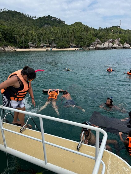 A group of people snorkeling in a clear blue sea, with a boat and a beach with palm trees in the background. 4