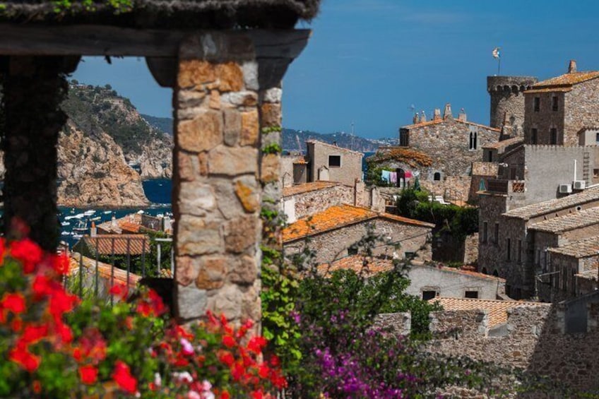 A coastal village with stone buildings, a stone archway, and red flowers in the foreground. 4