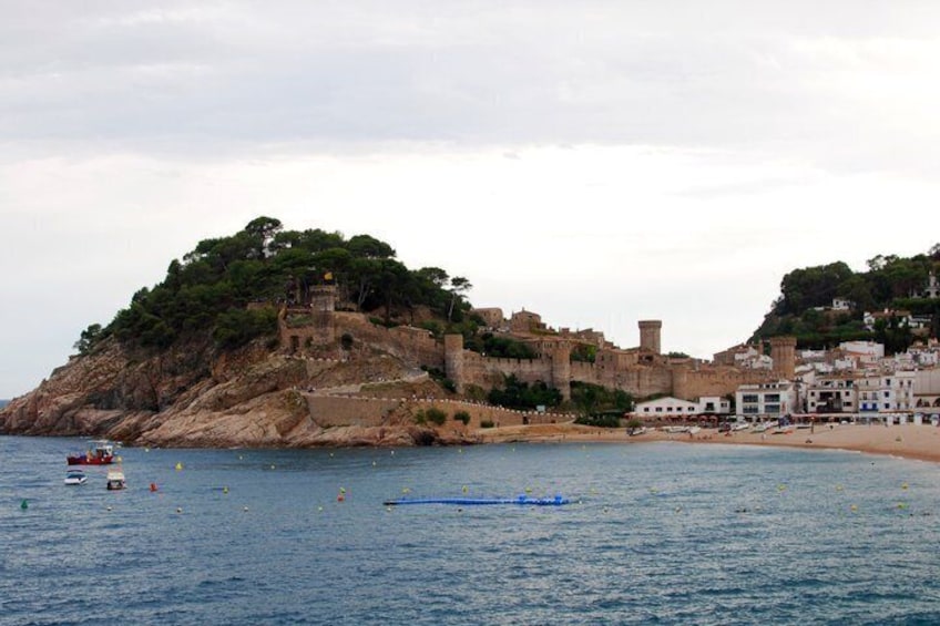 A coastal town with a historic castle on a hill, a beach, and calm sea. 2
