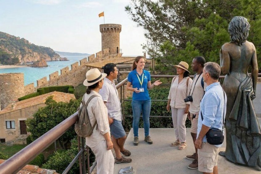 A group of people on a stone pathway with a coastal view, a statue, and a castle-like structure in the background. 1