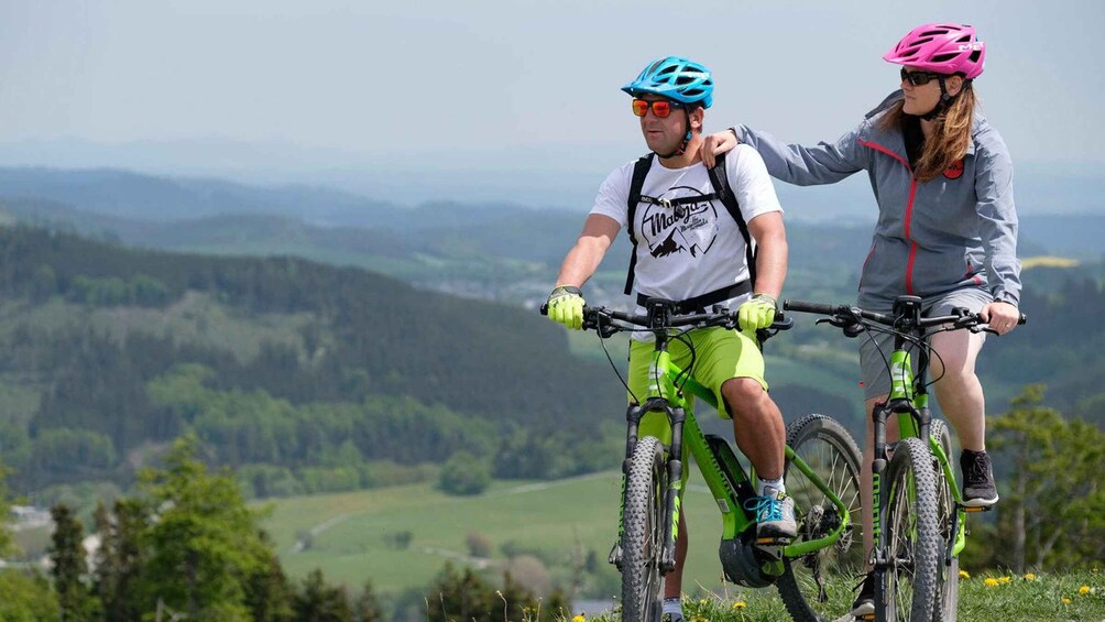 Two cyclists on a mountain trail with a scenic view of rolling hills and a valley. 1