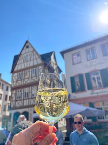 A hand holding a glass of white wine with 'Wiener' written on it, in front of a half-timbered building with a sign that reads 'Gasthof.' 3