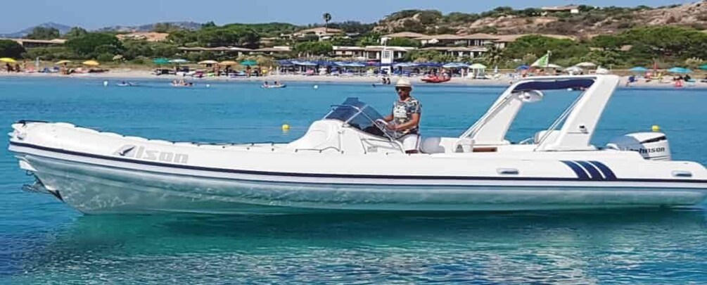 A white motorboat with 'Alson' written on the side, on calm blue water with a beach and houses in the background. 1