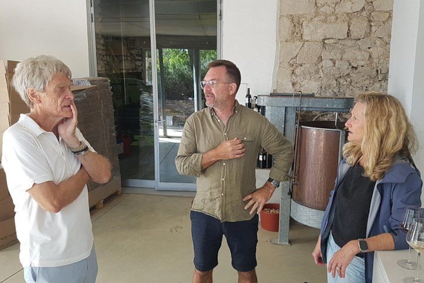 Three people standing indoors near a large copper vessel, possibly a distillation apparatus, with a stone wall and a glass door leading outside. 2
