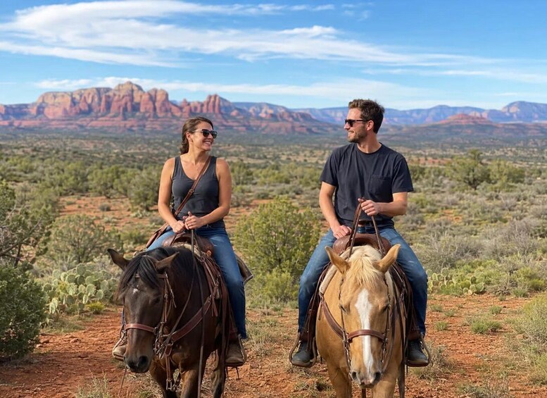 Two people riding horses on a trail with red rock formations in the background. 1