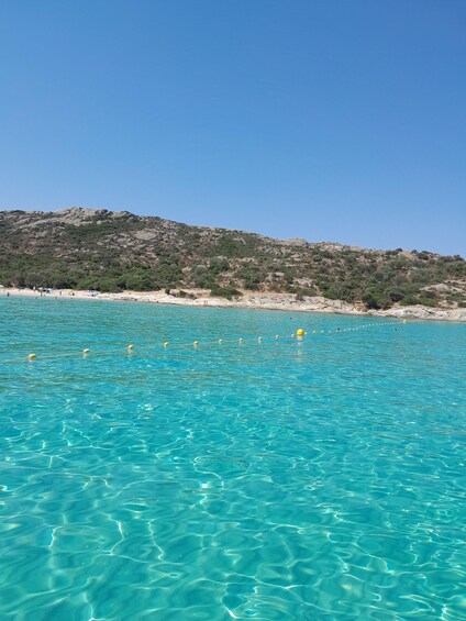A clear turquoise sea with buoys, a sandy beach, and a rocky hillside. 3