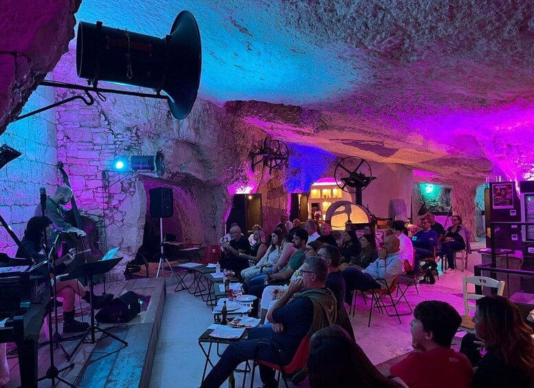 An indoor event with people seated in a room with a rustic stone wall, a stage with musical instruments, and colorful lighting. 4