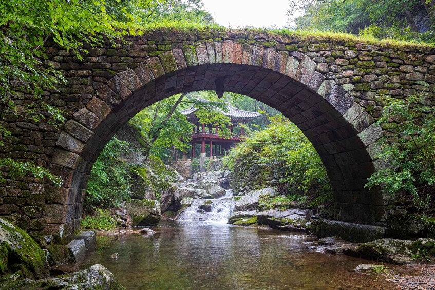 A stone bridge arching over a stream with a traditional building in the background. 1