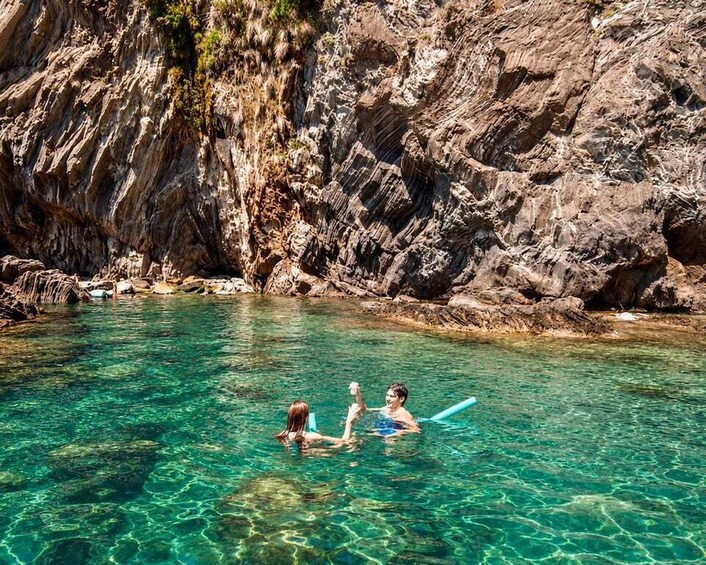 Two people swimming in a clear water body with rocky cliffs in the background. 1