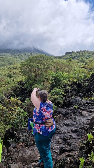 A person taking a photo of a volcanic landscape with lush greenery and clouds in the background. 5