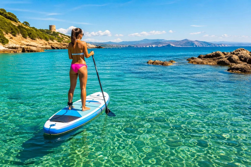 A person paddleboarding on clear turquoise water with a rocky coastline and a distant castle. 1