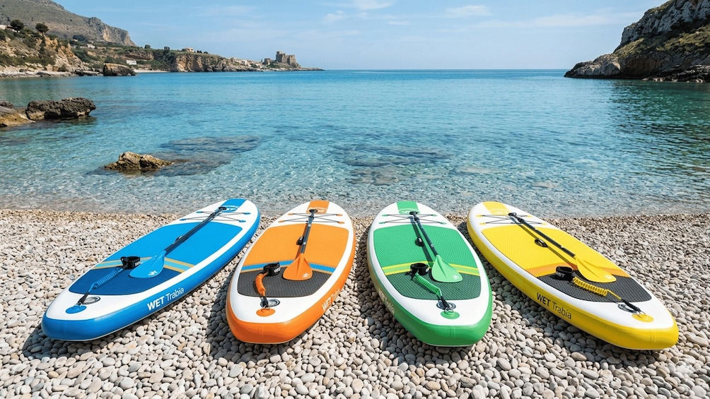 Four paddleboards on a pebbled beach with a clear blue sea and rocky cliffs in the background. 3
