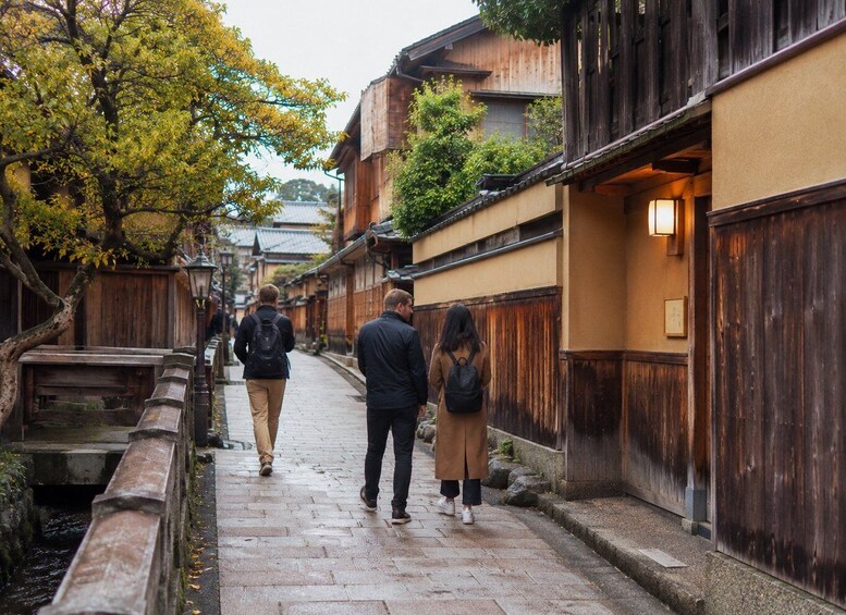 A narrow stone pathway flanked by traditional wooden buildings with wooden railings and a tree with yellow leaves. 5