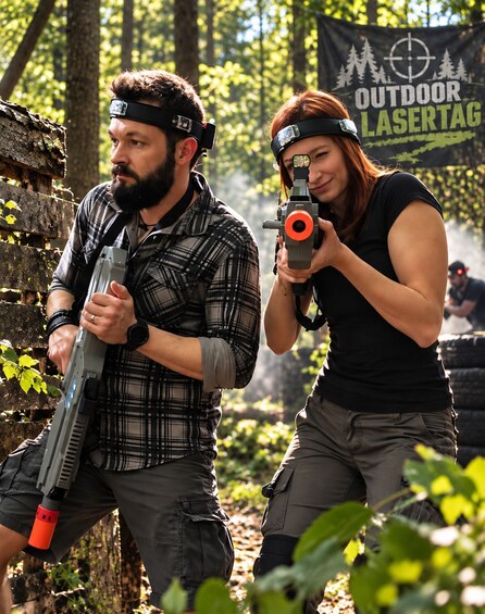 Two people in an outdoor setting, one holding a rifle and the other holding a pistol, with a banner in the background that reads 'OUTDOOR LASERTAG'. 1