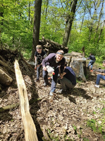 People in a forest clearing, some carrying tools, with a pile of logs and a large tree stump. 3