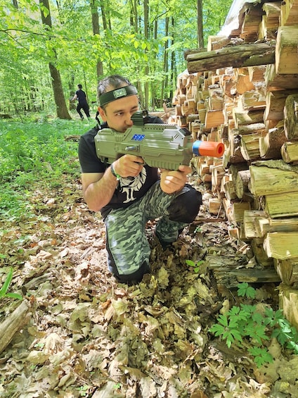 A person crouching in a forest holding a toy gun, surrounded by a stack of logs. 2