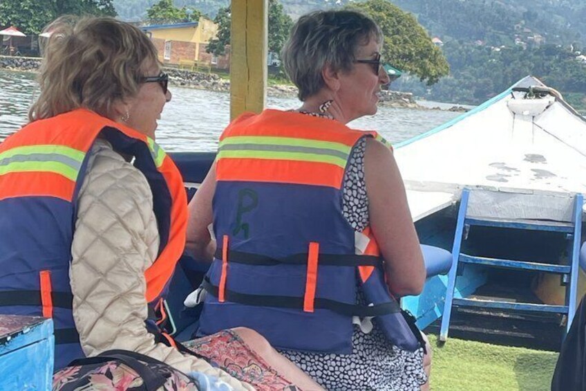 Two people wearing life jackets on a boat, with a dock and greenery in the background. 1
