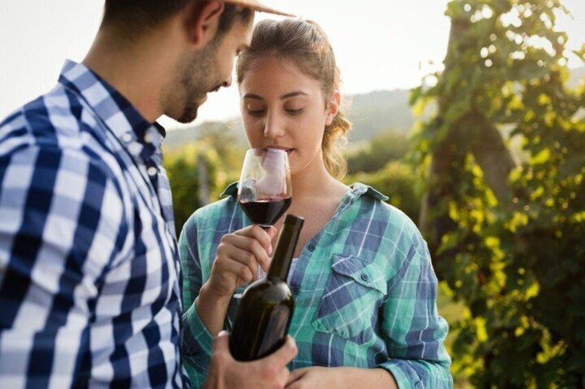 A person holding a wine bottle and glass, standing outdoors with greenery in the background. 1