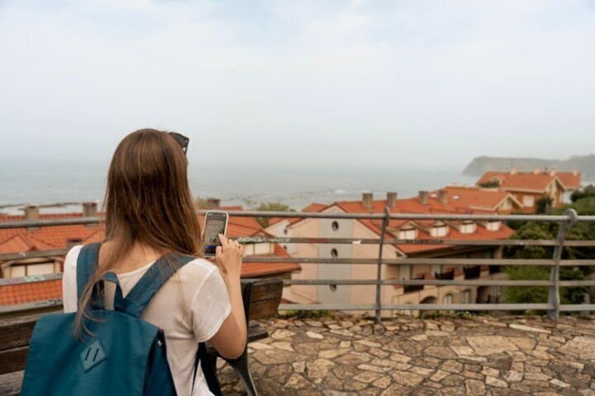 A person taking a photo with a smartphone, overlooking a coastal town with red-roofed buildings. 3