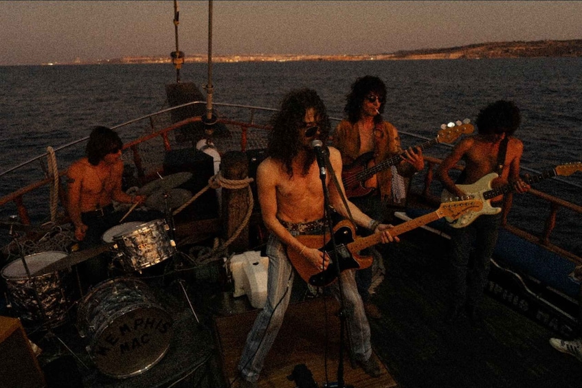 A band performing on a boat with a drum set and guitars, set against a backdrop of water and a distant shoreline. 4
