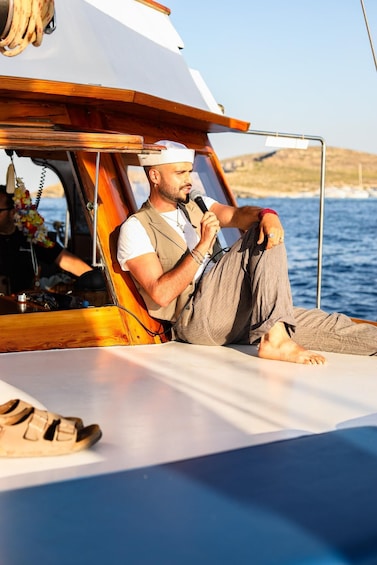 A person sitting on a boat, holding a microphone, with a calm sea and a hilly landscape in the background. 1