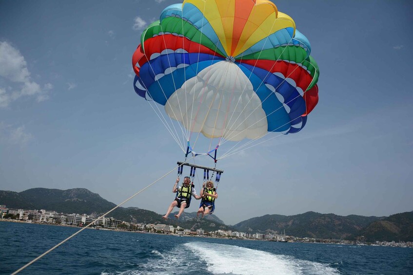 Two people parasailing over a body of water with a colorful parachute, with a coastal town and mountains in the background. 2