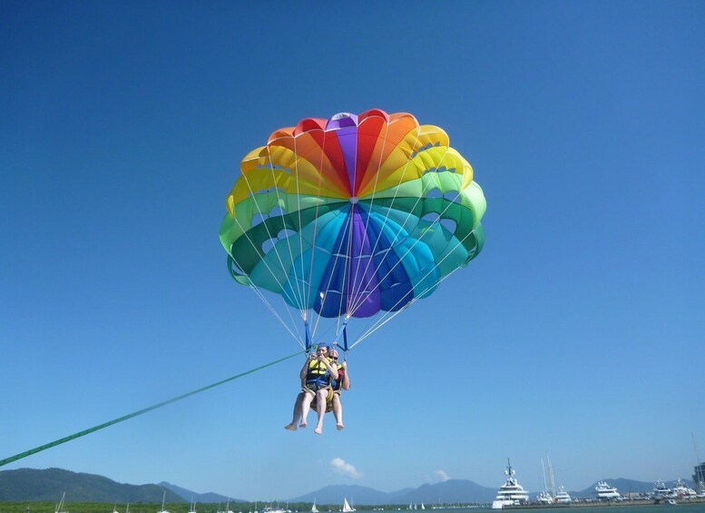 A person parasailing with a rainbow-colored parachute over a marina with boats and mountains in the background. 5