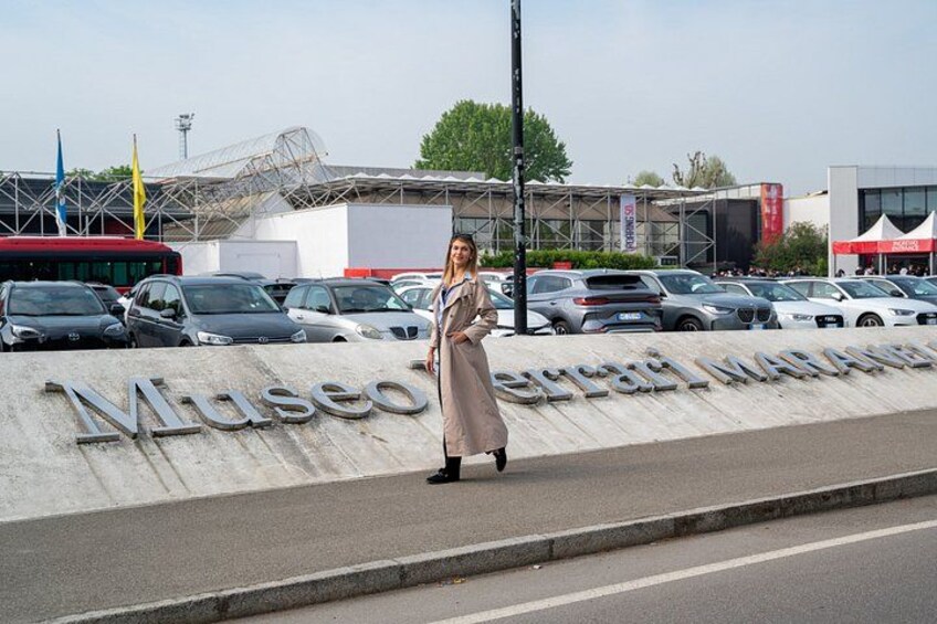 A woman walking past a sign that reads 'Museo Graciano Maranello' with a parking lot and cars in the background. 4