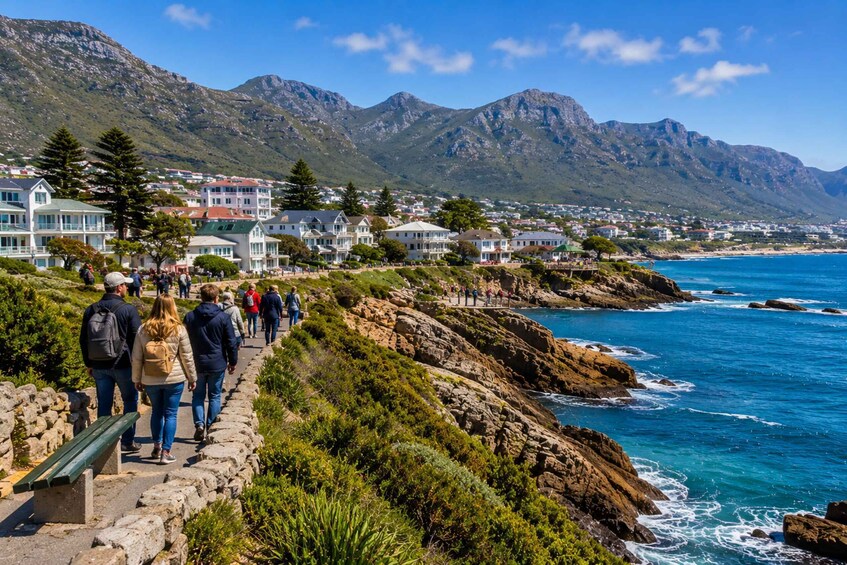 A coastal walkway with people walking, overlooking a rocky shoreline and a mountain range. 4
