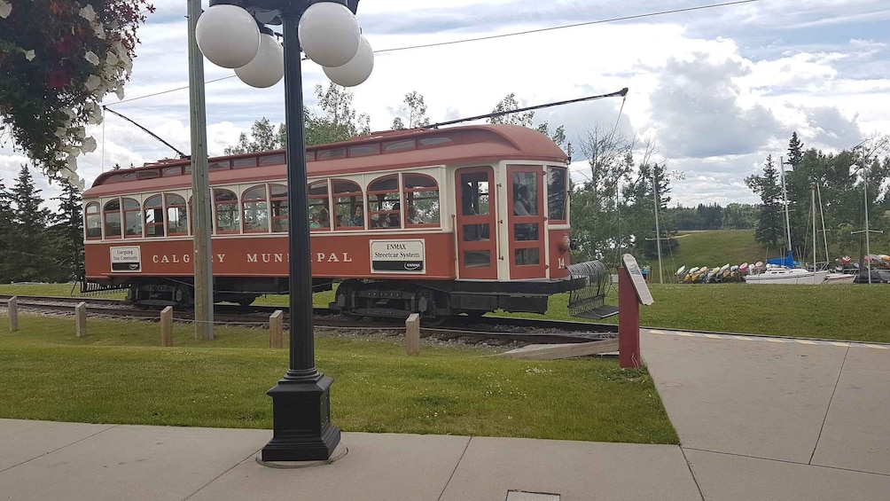 A red streetcar with 'CALGARY MUNIPAL' and 'ENMAX Streetcar System' on its side, parked on tracks with a grassy area and trees in the background. 2