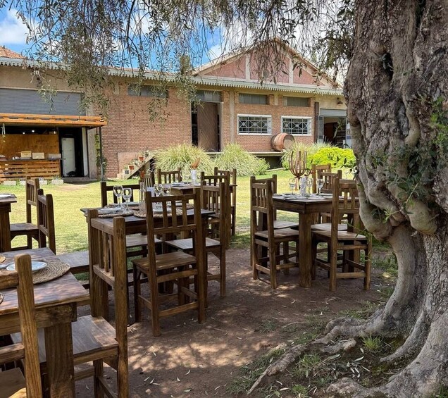 An outdoor dining area with wooden tables and chairs, surrounded by a garden and a brick building. 1