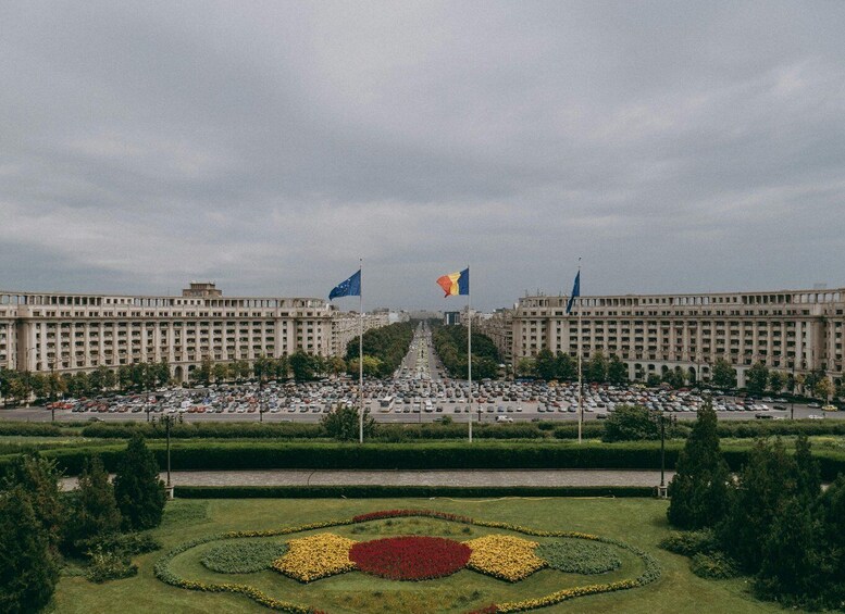 A large building with multiple flags, a busy street, and a well-maintained garden with geometric flower beds. 5