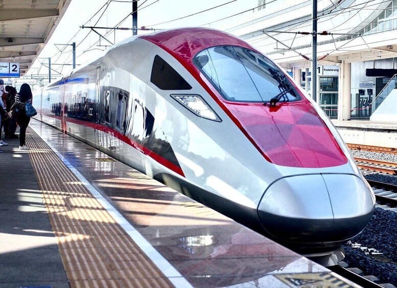 A high-speed train at a station platform with overhead electrical lines and a pedestrian walkway. 1