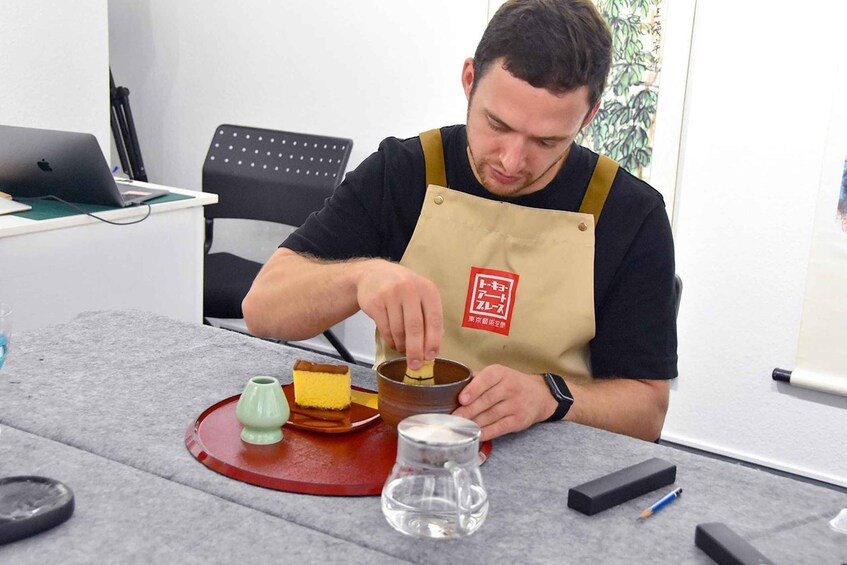 A person wearing a brown apron with a logo, stirring a liquid in a metal container on a table with a red mat. 2