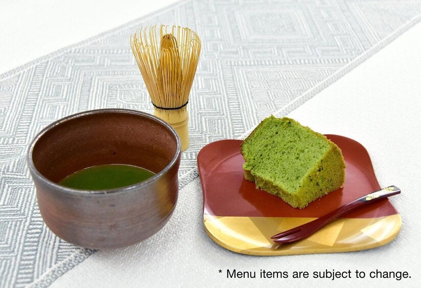 A green cake on a red plate with a whisk and a cup of green tea on a patterned tablecloth. 5