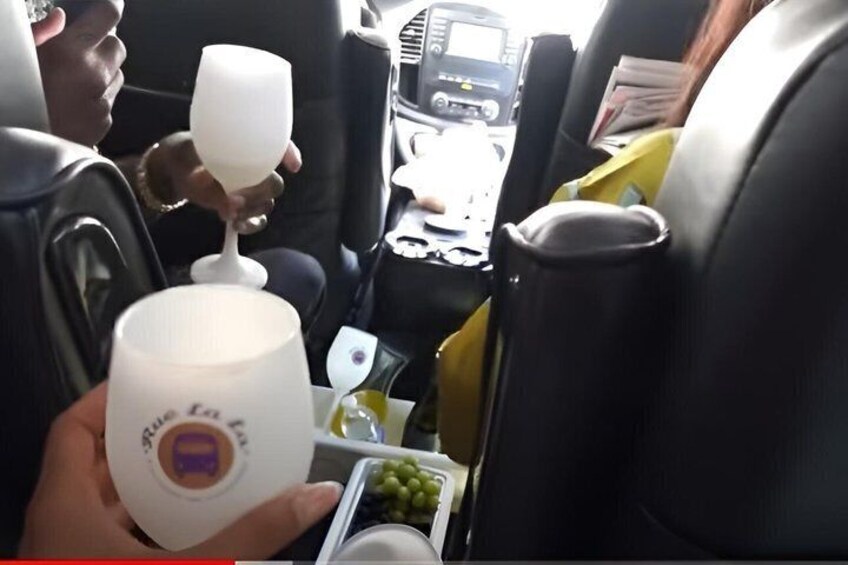 A person holding a white cup with a purple logo, a car interior with black seats, and a cup holder with a bottle and grapes. 5