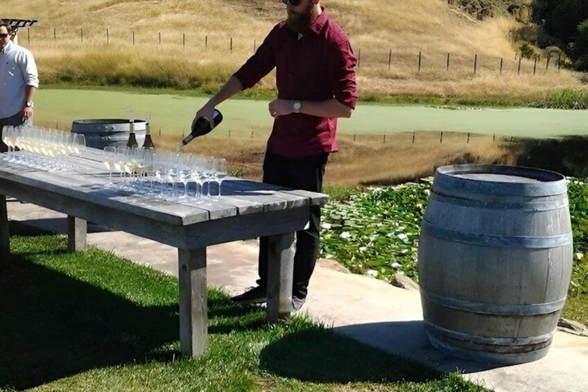 A person pouring liquid into glasses on a table outdoors with a large barrel nearby. 4