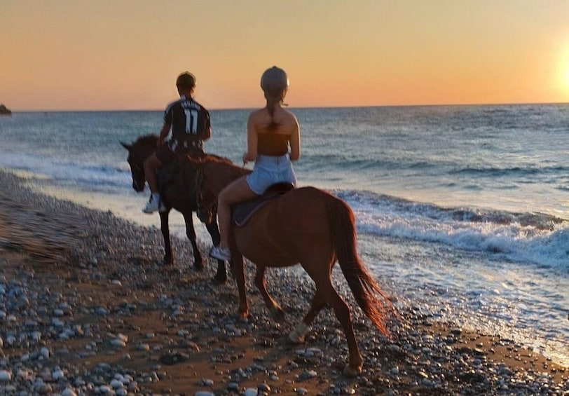 Two people riding horses along a pebbled beach at sunset. 3