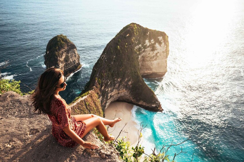 A person sitting on a rock by the ocean, overlooking a beach with large rock formations. 5