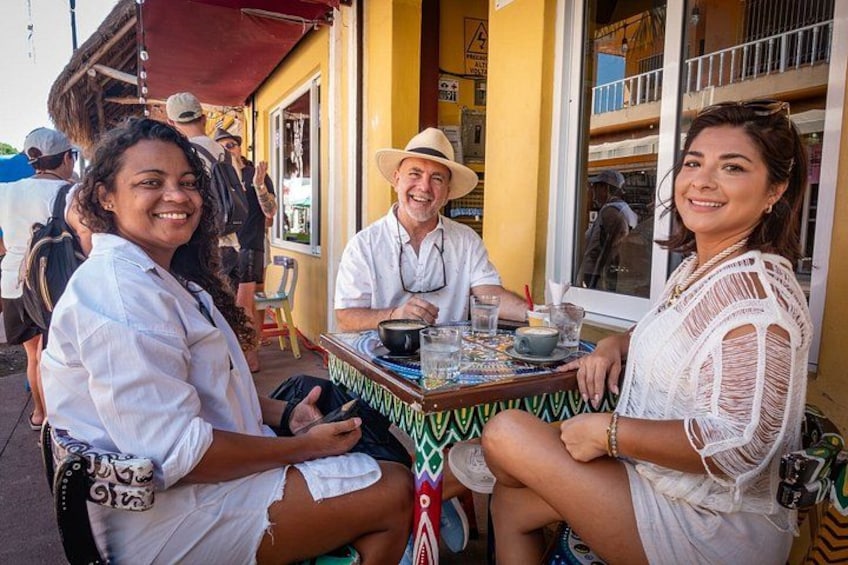A group of people sitting at an outdoor table with a colorful tablecloth, a yellow building in the background, and a red awning. 5