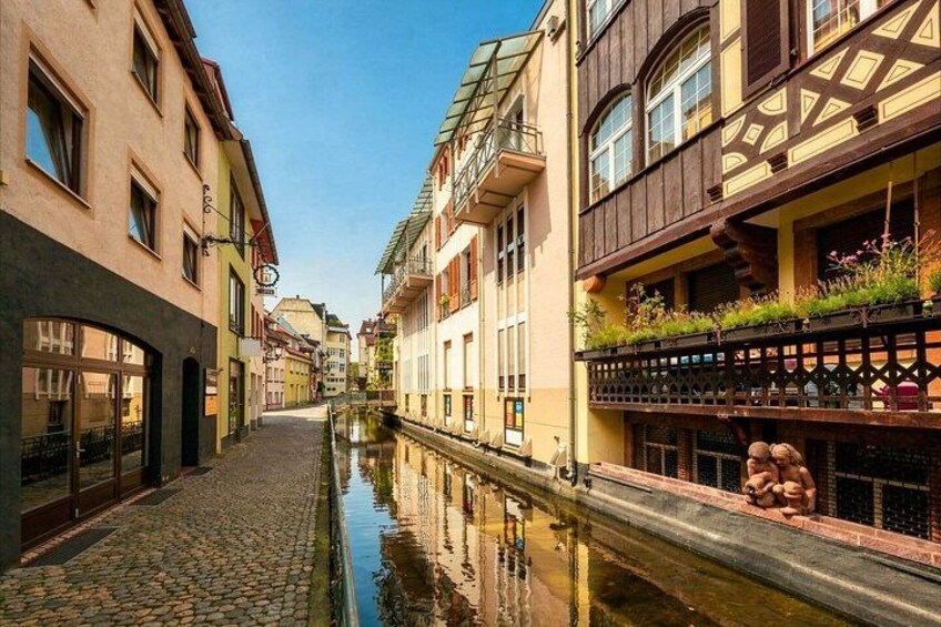 A cobblestone street with buildings on either side, a canal, and a bridge with people on it. 4