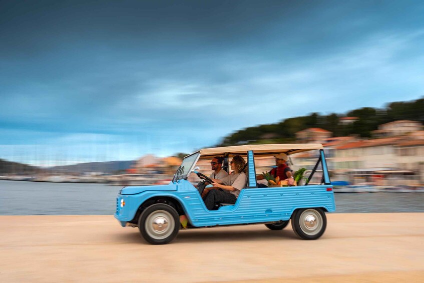A blue vintage car with passengers driving along a waterfront. 3