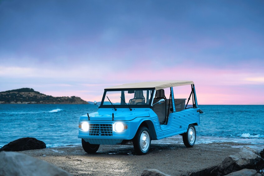 A blue vintage car parked on a rocky beach with the ocean and a sunset in the background. 2