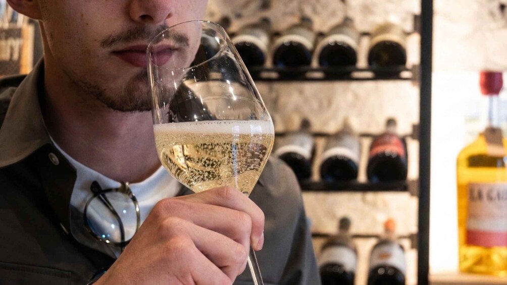 Une personne tenant un verre de vin blanc, debout dans une cave à vin, avec des bouteilles en arrière-plan. 2