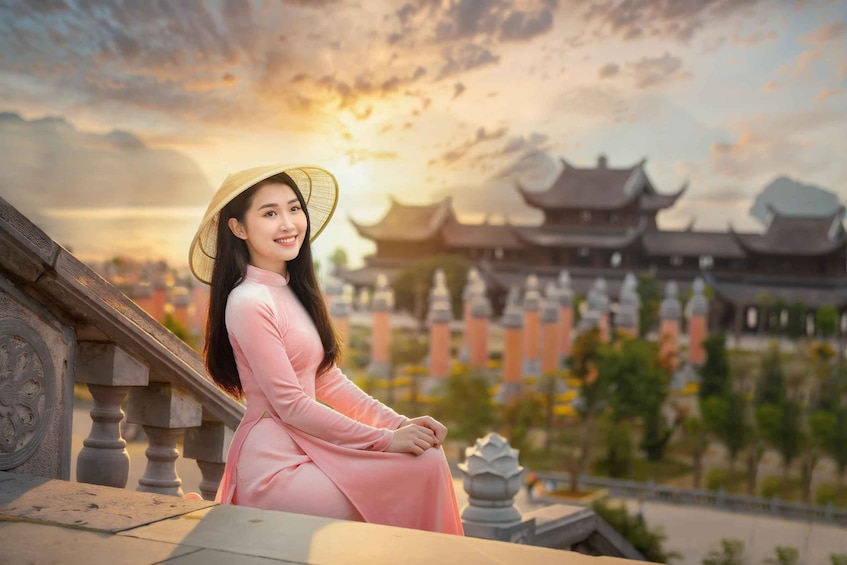 A person in a pink dress and straw hat sitting on a stone balustrade with traditional architecture in the background. 4