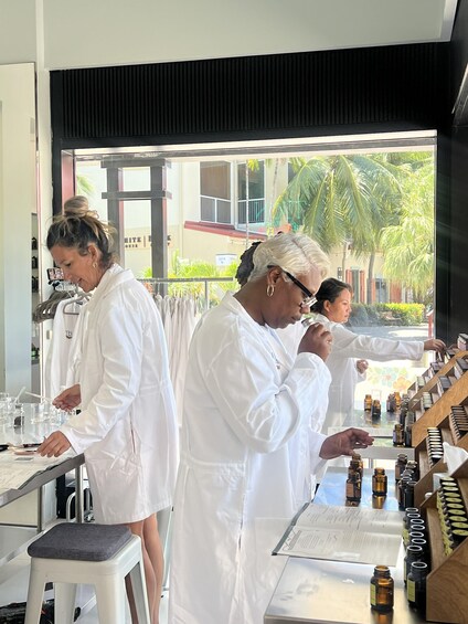 Three people in white lab coats at a counter with various bottles, in a room with a view of palm trees and a building outside. 5