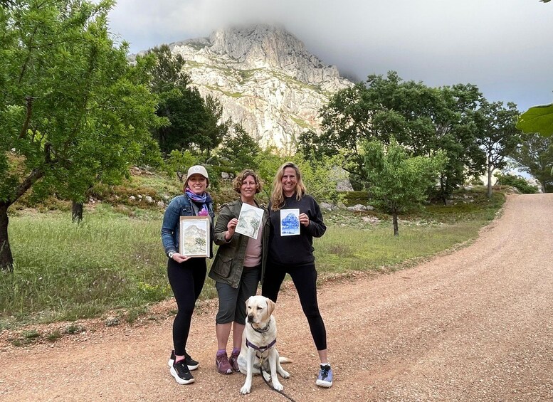 Three people holding framed pictures, a dog, and a mountain in the background. 1
