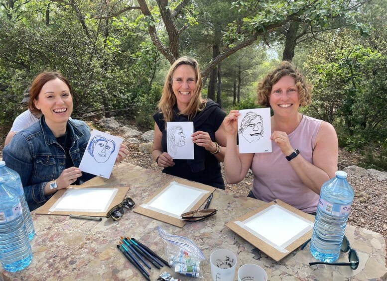 Three people holding drawings at an outdoor table with art supplies. 3