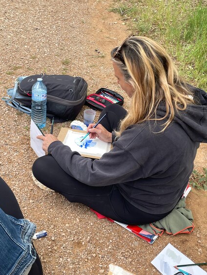 A person sitting on the ground, sketching on paper with a pencil, surrounded by art supplies and a water bottle. 2